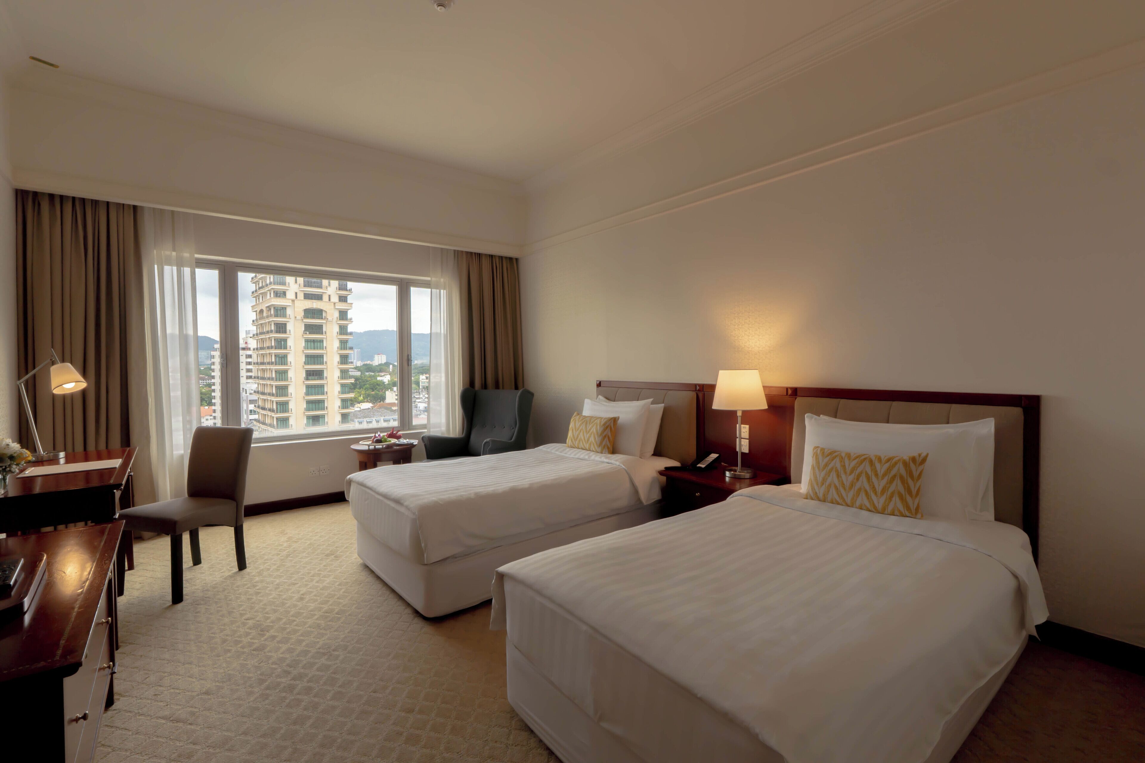 A hotel room with two beds, a desk, a chair, a lamp, and a view of a cityscape through the window.