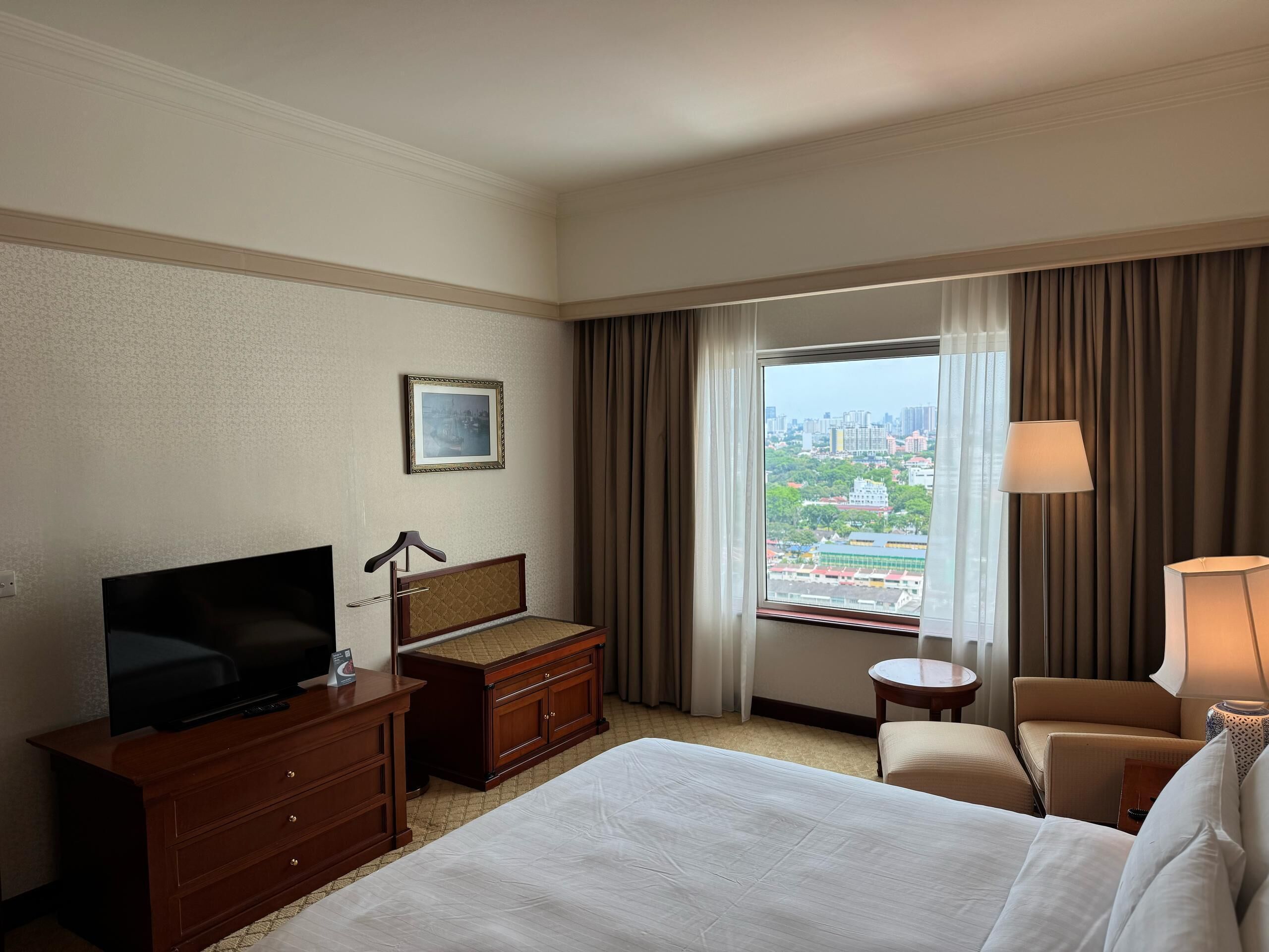 A hotel room with a bed, a TV, a dresser, a desk, a chair, a lamp, and a window with a city view.