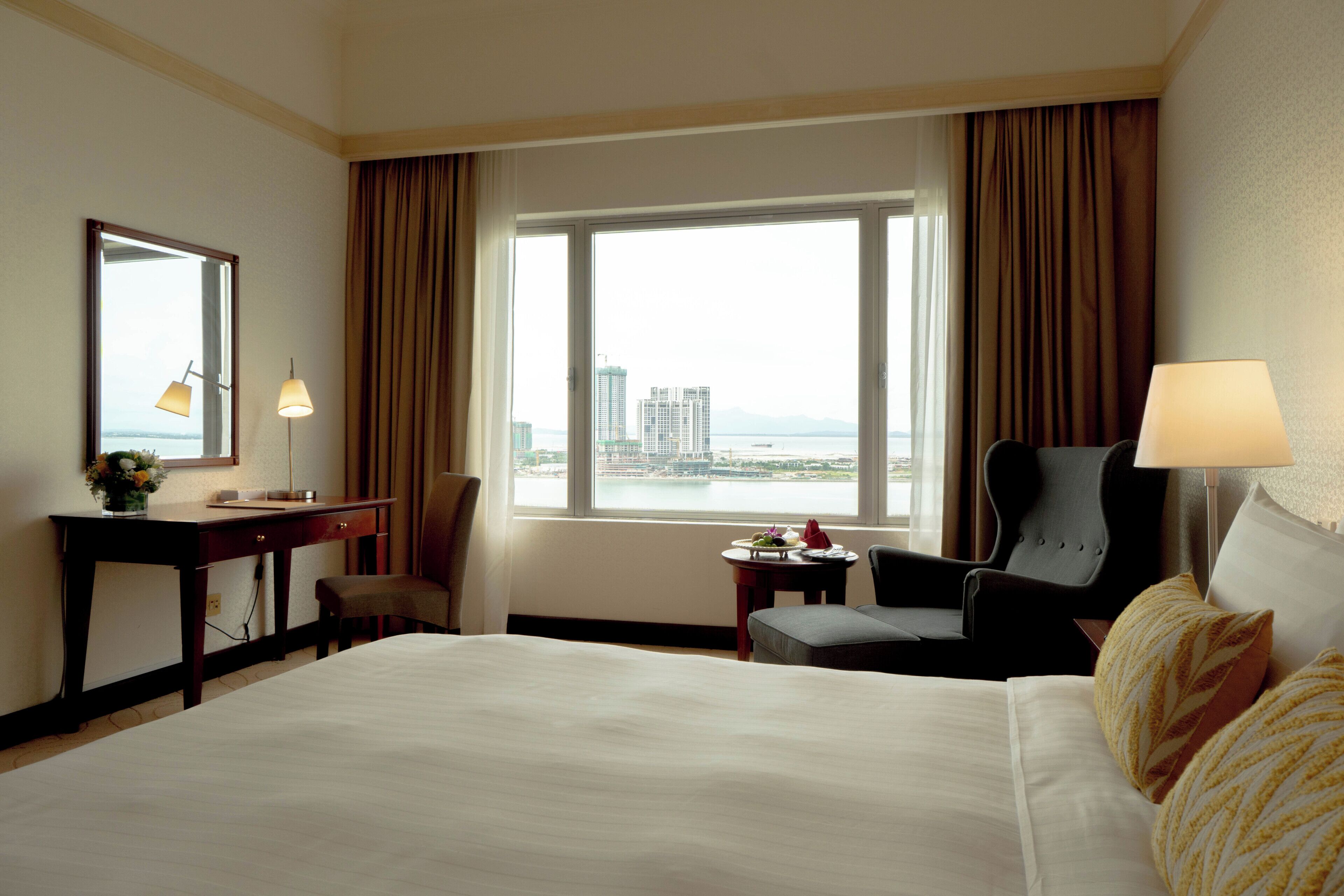 A hotel room with a large bed, a desk, a chair, a small table with a tray of fruit, and a view of a cityscape through the window.