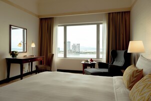 A hotel room with a large bed, a desk, a chair, a small table with a tray of fruit, and a view of a cityscape through the window.