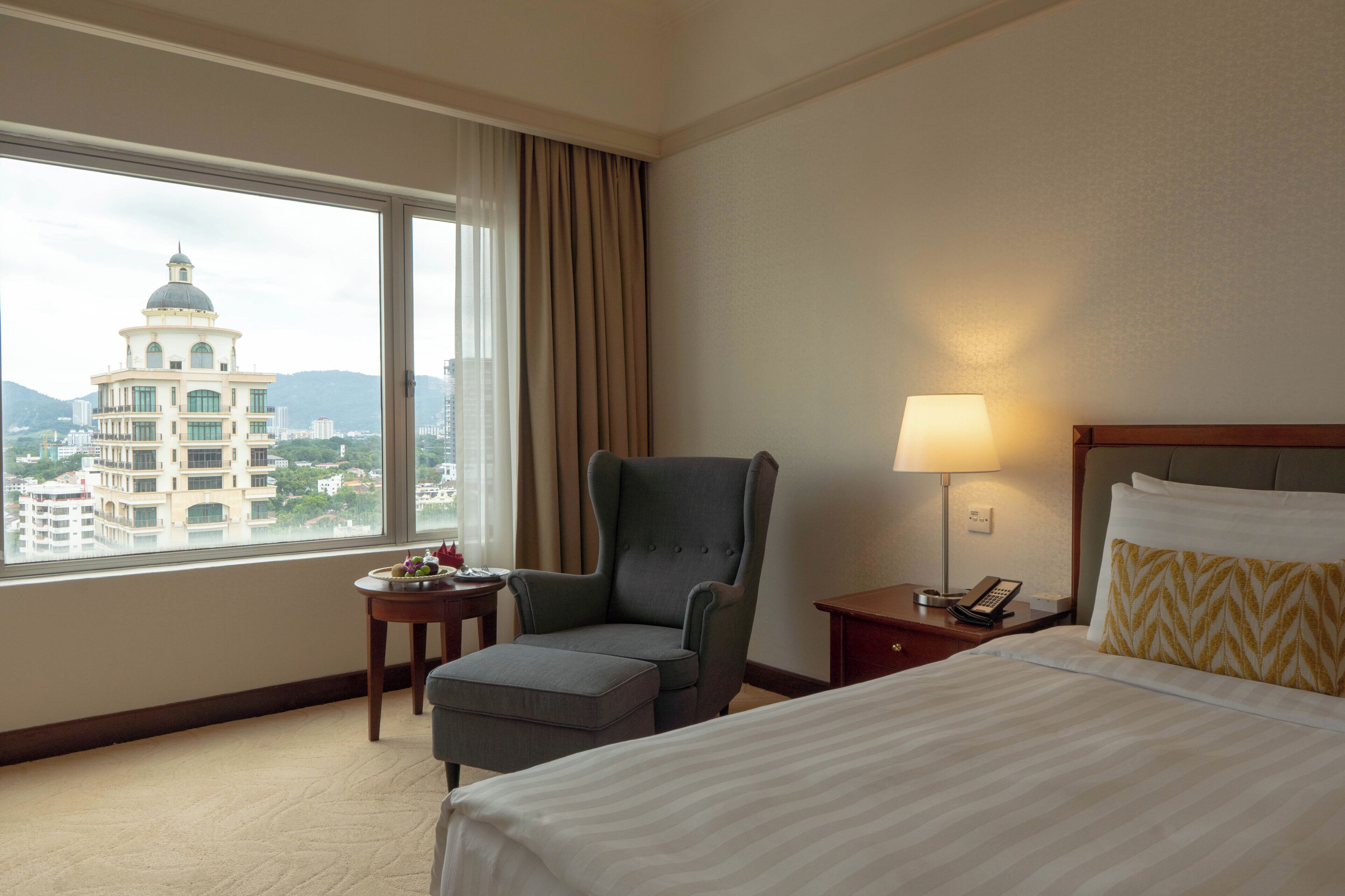 A hotel room with a bed, a chair, a small table, a lamp, and a view of a cityscape with a building with a dome.