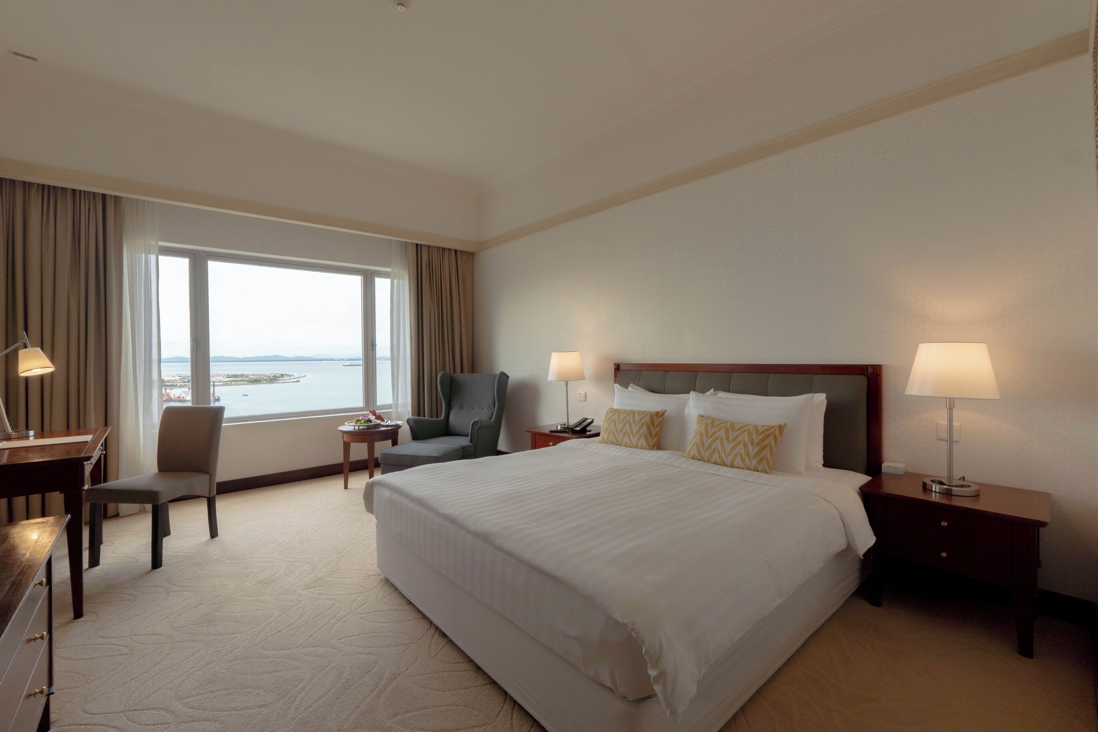 Une chambre d’hôtel avec un grand lit, un bureau, une chaise, une table de chevet et une vue sur la mer.