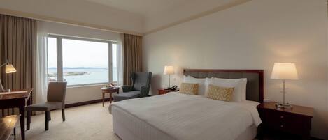 A hotel room with a large bed, a desk, a chair, a nightstand, and a view of the sea.