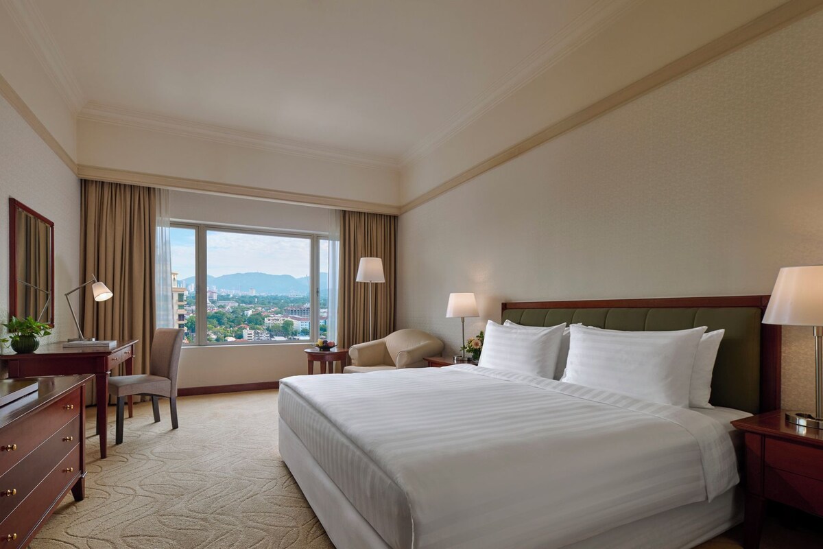 A hotel room with a large bed, a desk with a computer, a chair, a lamp, and a view of the city through the window.