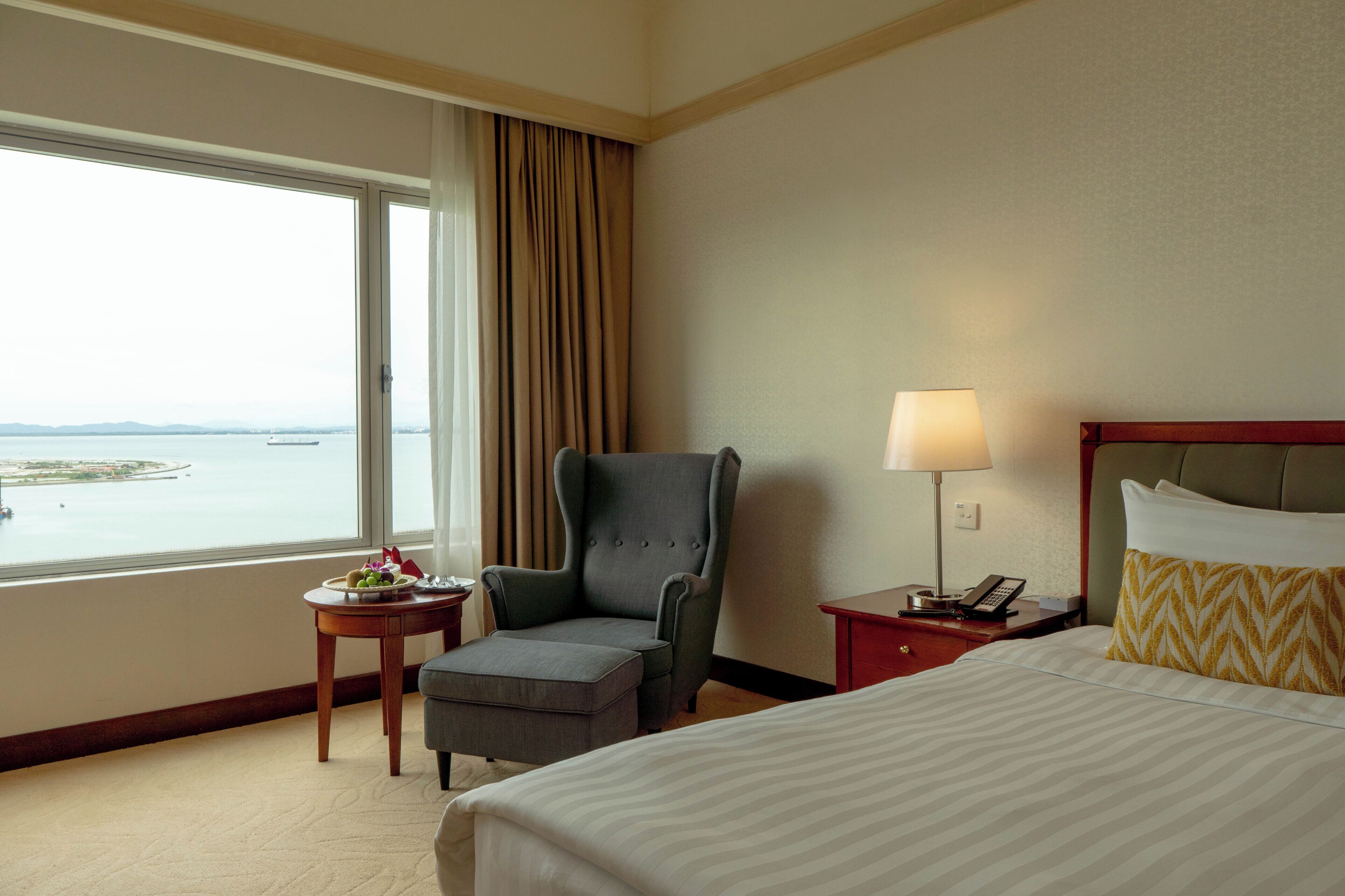A hotel room with a bed, a chair, a small table with a fruit bowl, a lamp, and a view of the sea.