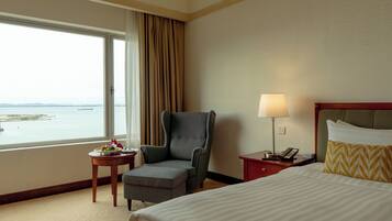 A hotel room with a bed, a chair, a small table with a fruit bowl, a lamp, and a view of the sea.