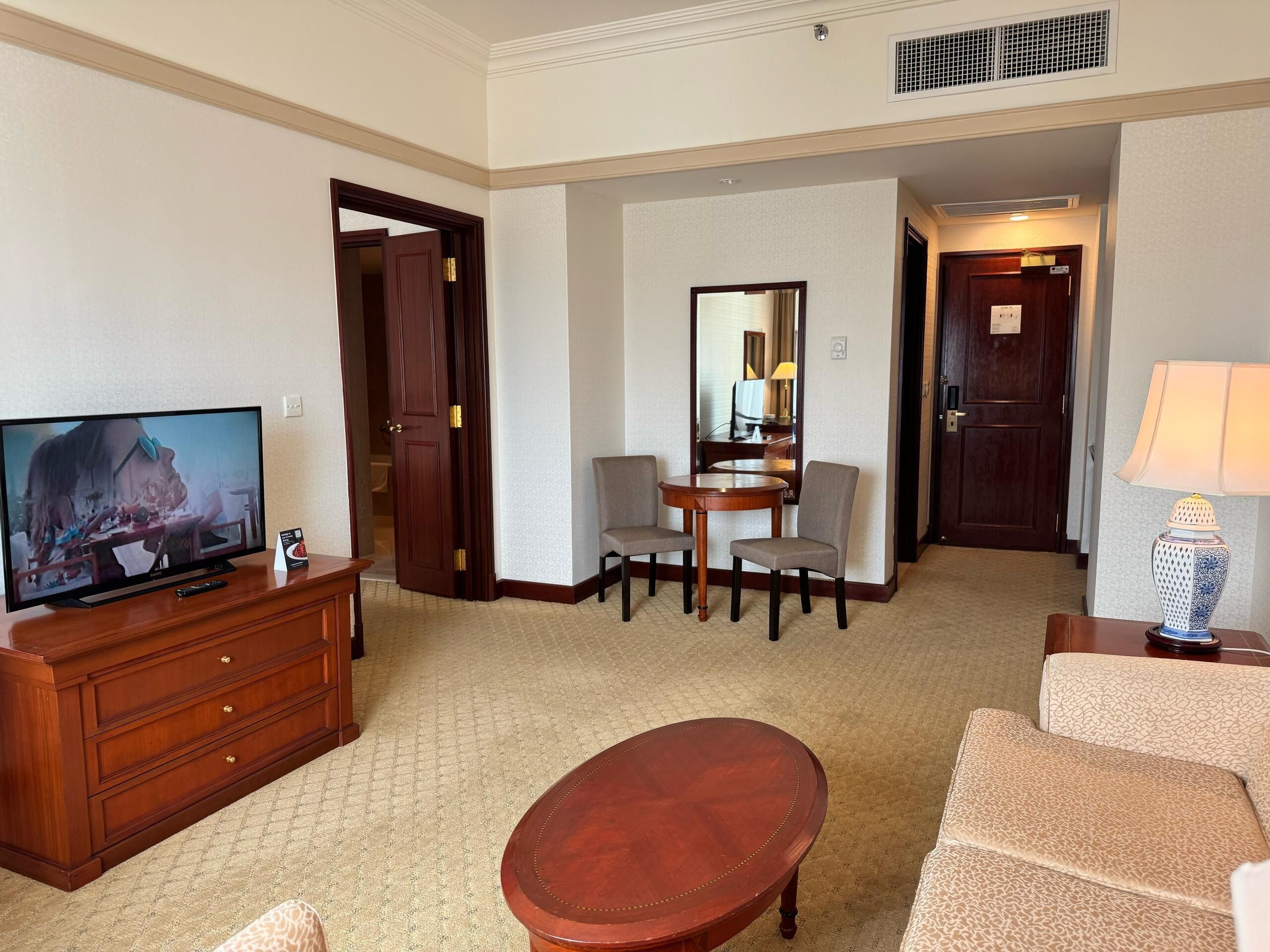 A hotel room with a television, a wooden dresser, a round coffee table, a sofa, a dining table with chairs, and a mirror.