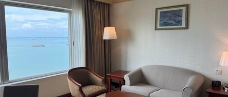 Habitación de hotel con un ventanal que ofrece vistas al mar, un sofá, una silla, una mesa y una lámpara.