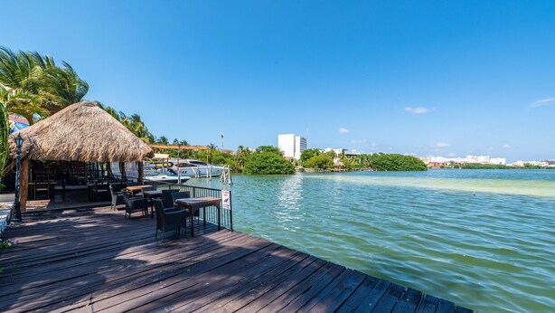 A wooden deck with tables and chairs, a thatched-roof structure, and a view of a calm body of water with buildings in the background. - Hotel Imperial Laguna Faranda (Cancun)
