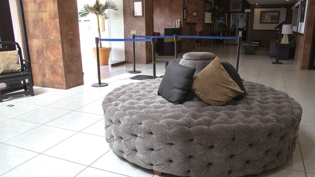 A round, tufted grey ottoman with multiple cushions in a lobby area with tiled flooring, a reception desk, and a blue rope barrier.