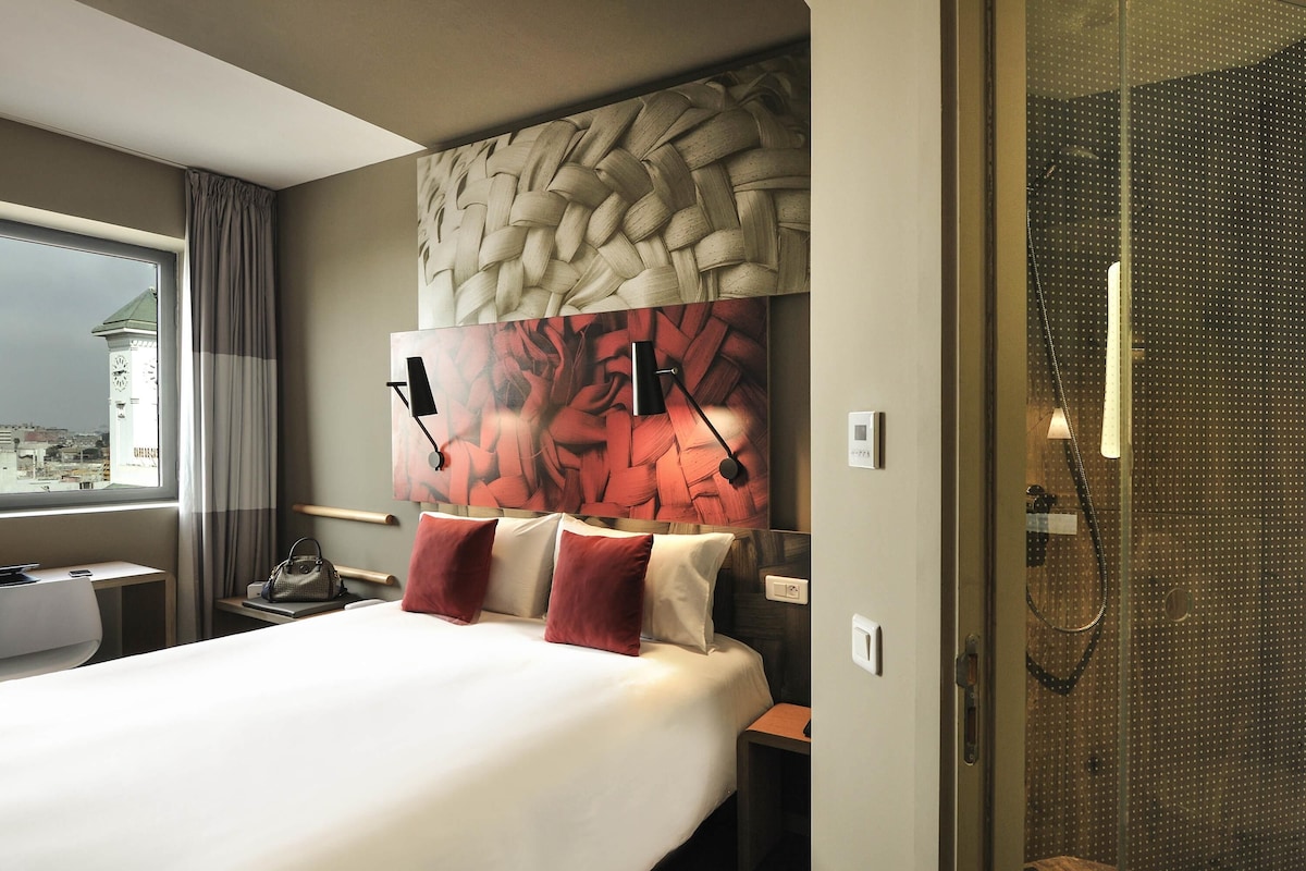 Habitación de hotel con una cama grande, cabecera con diseño tejido, mesita de noche, lámpara y vista a la ciudad a través de una ventana.