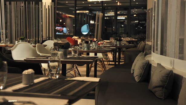 Un intérieur de restaurant moderne avec des tables et des chaises, un canapé avec des coussins et une grande fenêtre donnant sur la ville.
