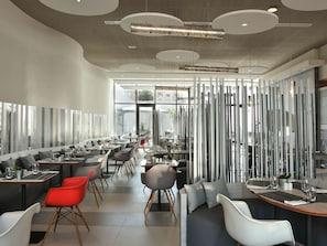 Un intérieur de restaurant moderne, avec de hauts plafonds, des luminaires suspendus et une vue dégagée grâce à de grandes fenêtres.