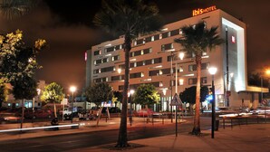 Un hôtel à plusieurs étages, doté d’une façade moderne, de fenêtres illuminées et d’une rue bordée de palmiers.