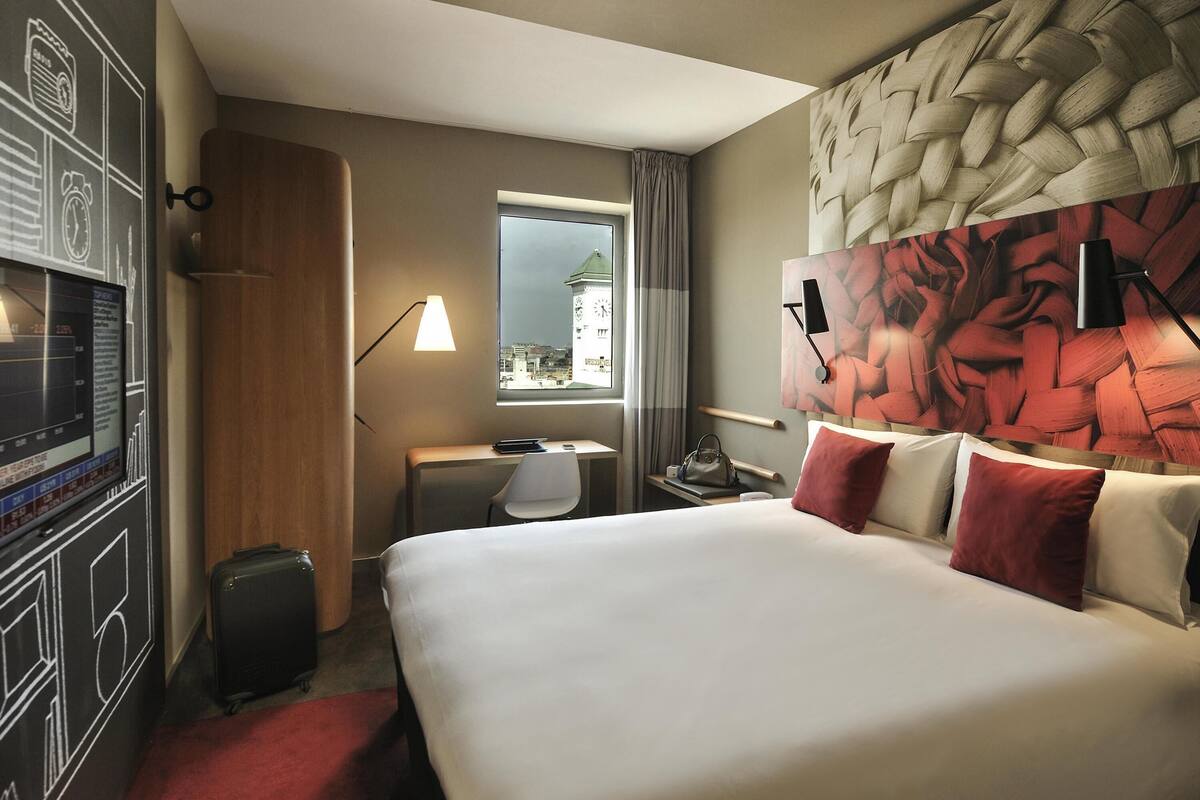 Een hotelkamer met een bed, een bureau, een stoel, een koffer, een televisie aan de muur en een raam met gordijnen.