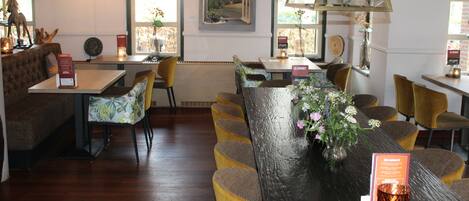 A restaurant with wooden tables and chairs, a large window with a painting, and pendant lighting.