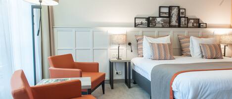 Quarto de hotel com uma cama, duas cadeiras laranja, uma mesinha, uma mesa de cabeceira e um abajur.