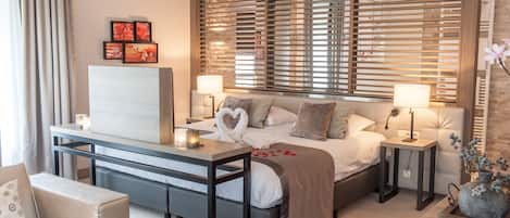 Una habitación de hotel moderna con una cama, mesitas de noche, un escritorio, un sofá y un amplio ventanal con persianas.