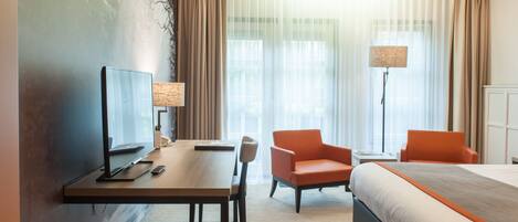 A hotel room with a bed, a desk with a computer, a chair, a lamp, and a large window with curtains.