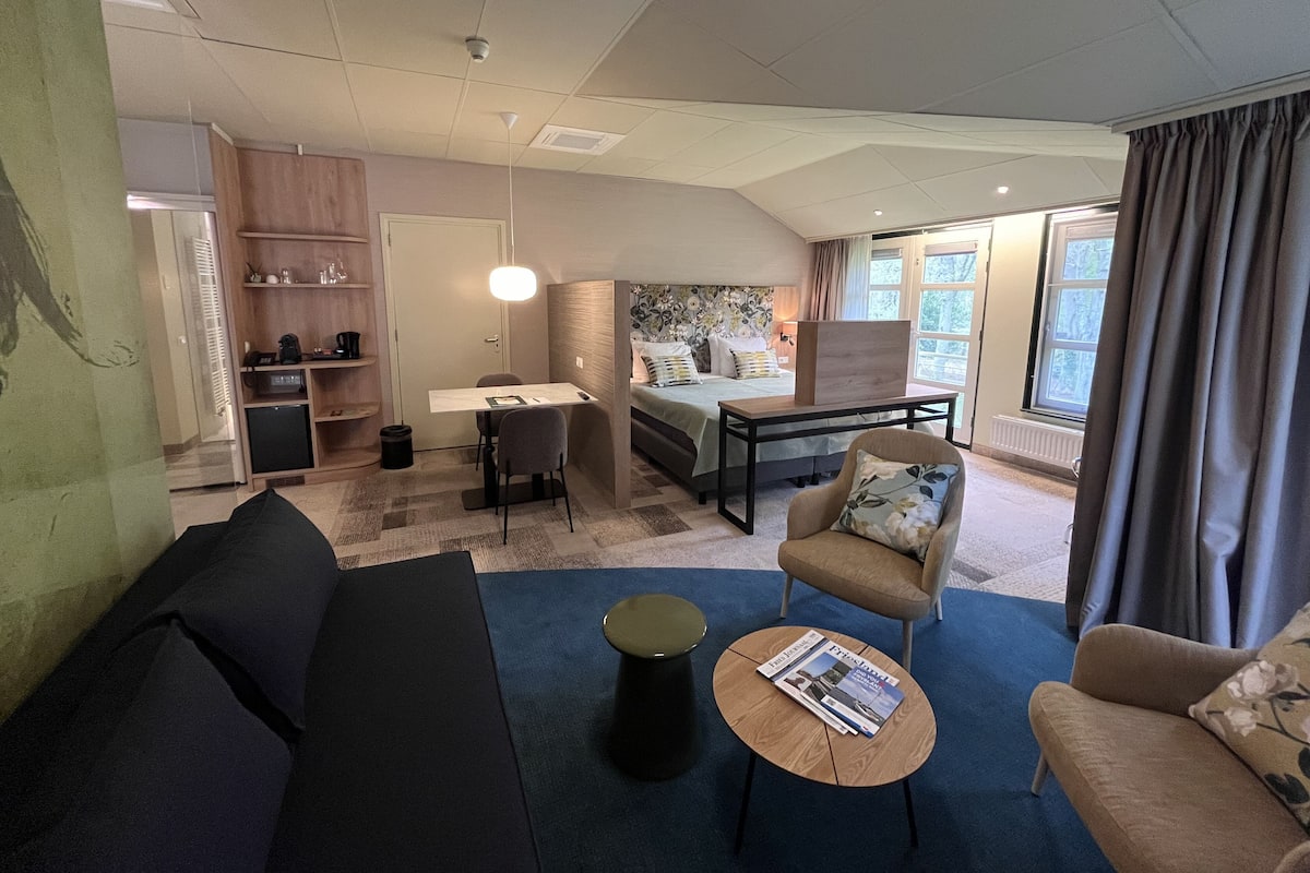 Une chambre d’hôtel moderne, équipée d’un canapé, d’un fauteuil, d’une table basse et d’un lit avec une tête de lit. On y trouve également un bureau avec une chaise, une bibliothèque et une fenêtre avec des rideaux.
