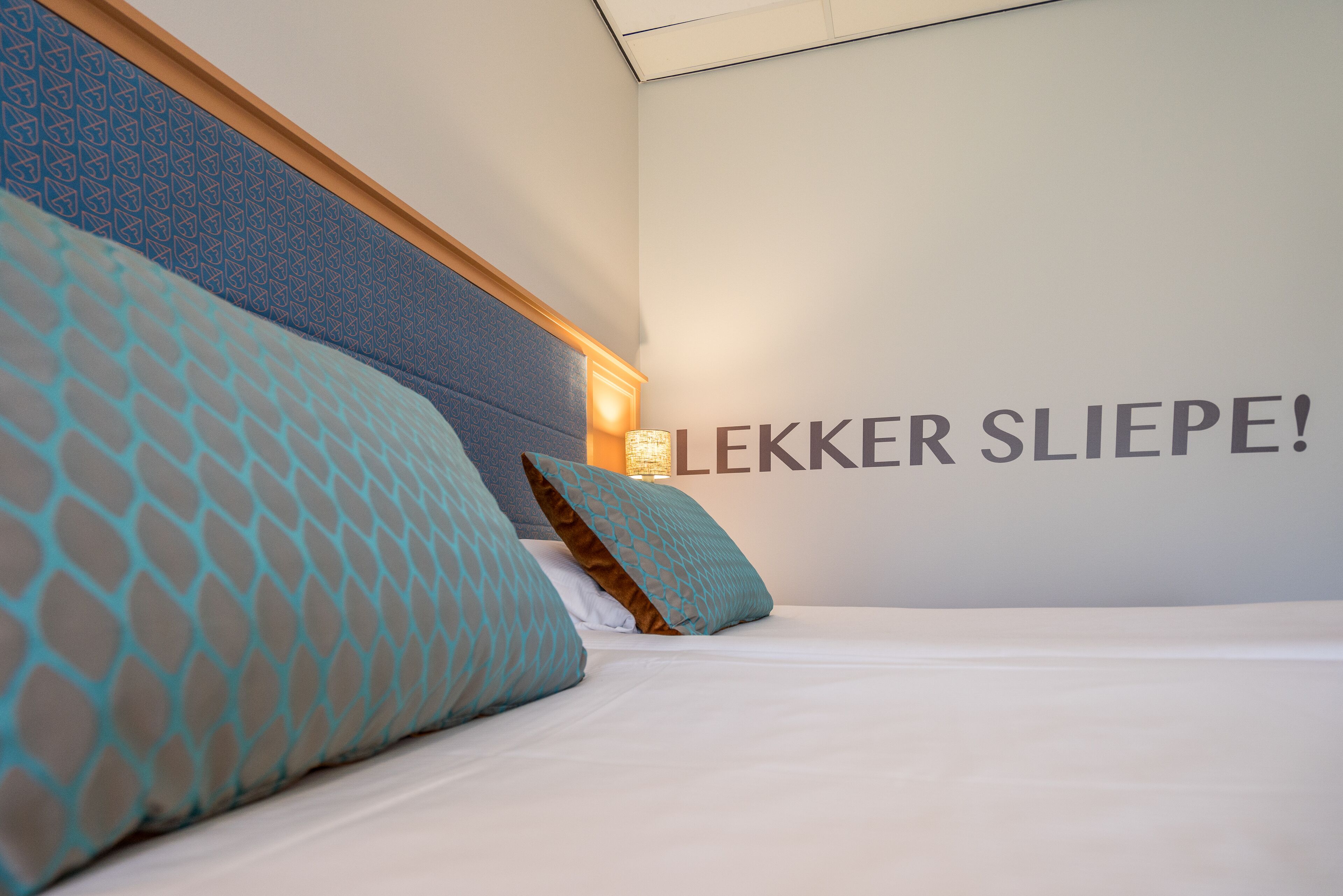 A hotel room with a bed, two pillows, a headboard, and a wall with the text "LEKKER SLIEPE!".
