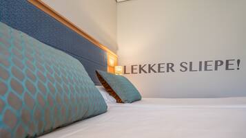 A hotel room with a bed, two pillows, a headboard, and a wall with the text "LEKKER SLIEPE!".
