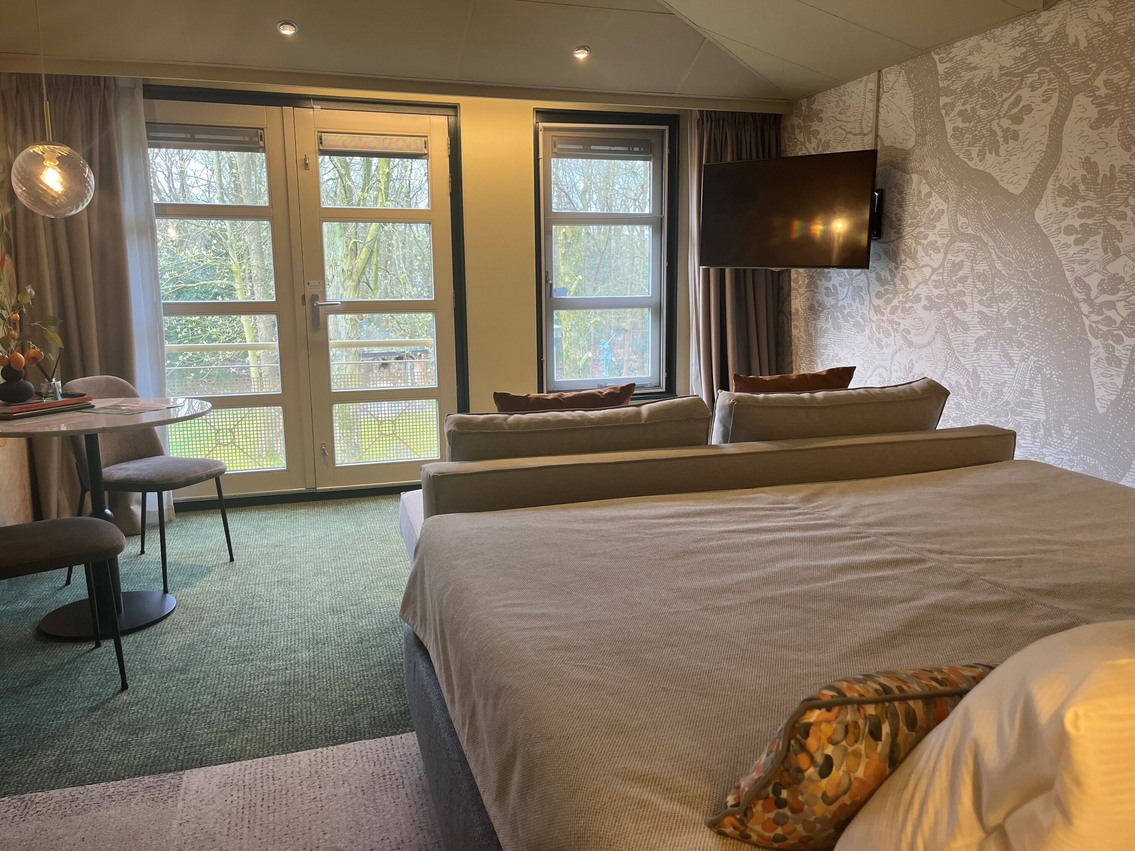 A hotel room with a bed, a desk, and a chair. There is a TV mounted on the wall and a window with curtains.