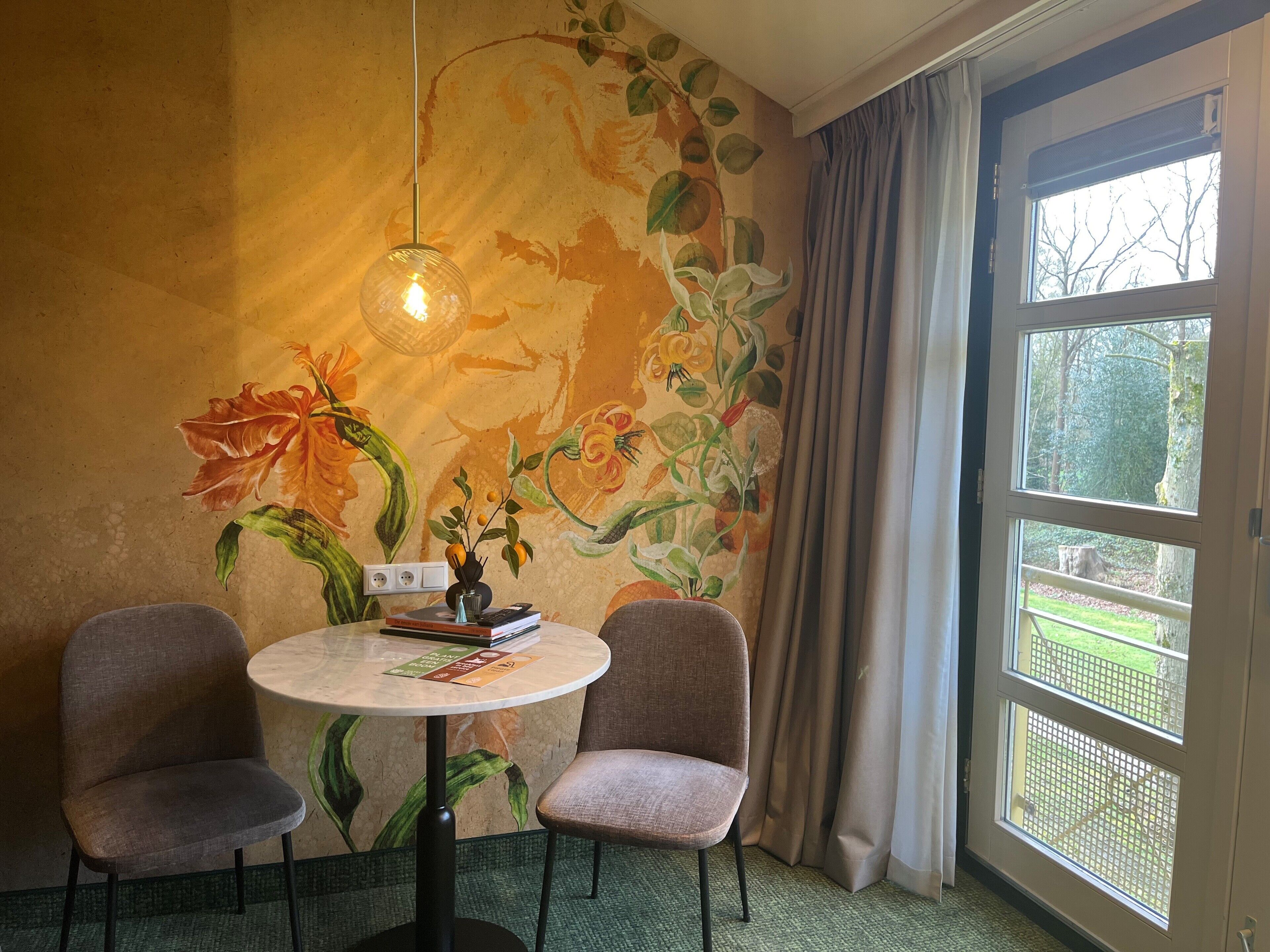 A room with a round table, two chairs, a floral mural, and a window with a view of greenery.