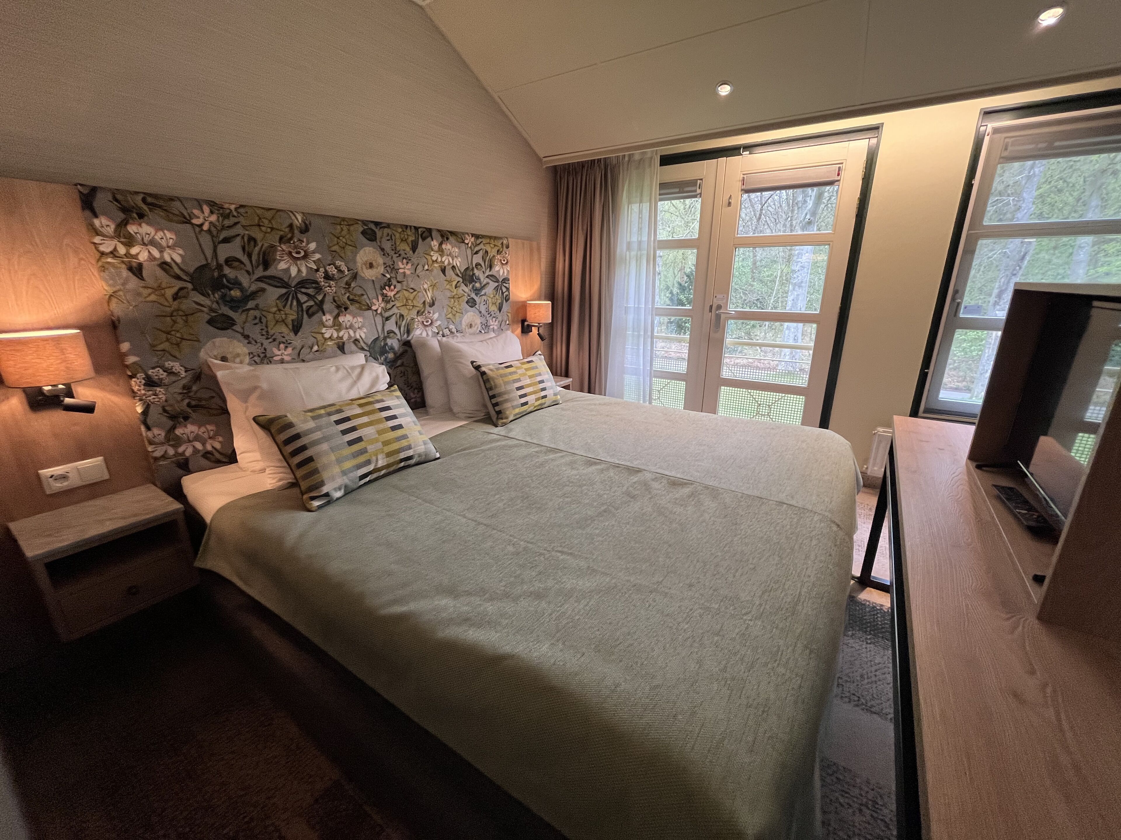 A hotel room with a large bed, two bedside tables, a TV, and a window with a view of trees.