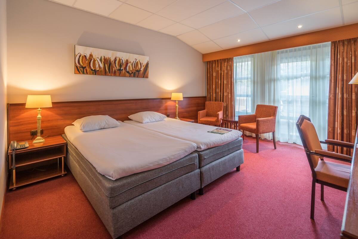 Une chambre d’hôtel avec un lit double, deux lampes de chevet, un bureau avec une chaise, une grande fenêtre avec des rideaux et un tableau au mur.