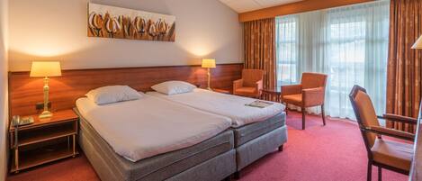 Une chambre d’hôtel avec un lit double, deux lampes de chevet, un bureau avec une chaise, une grande fenêtre avec des rideaux et un tableau au mur.