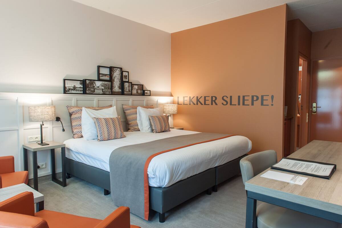 Une chambre d’hôtel moderne avec un grand lit, des tables de chevet, un bureau et des chaises orange.
