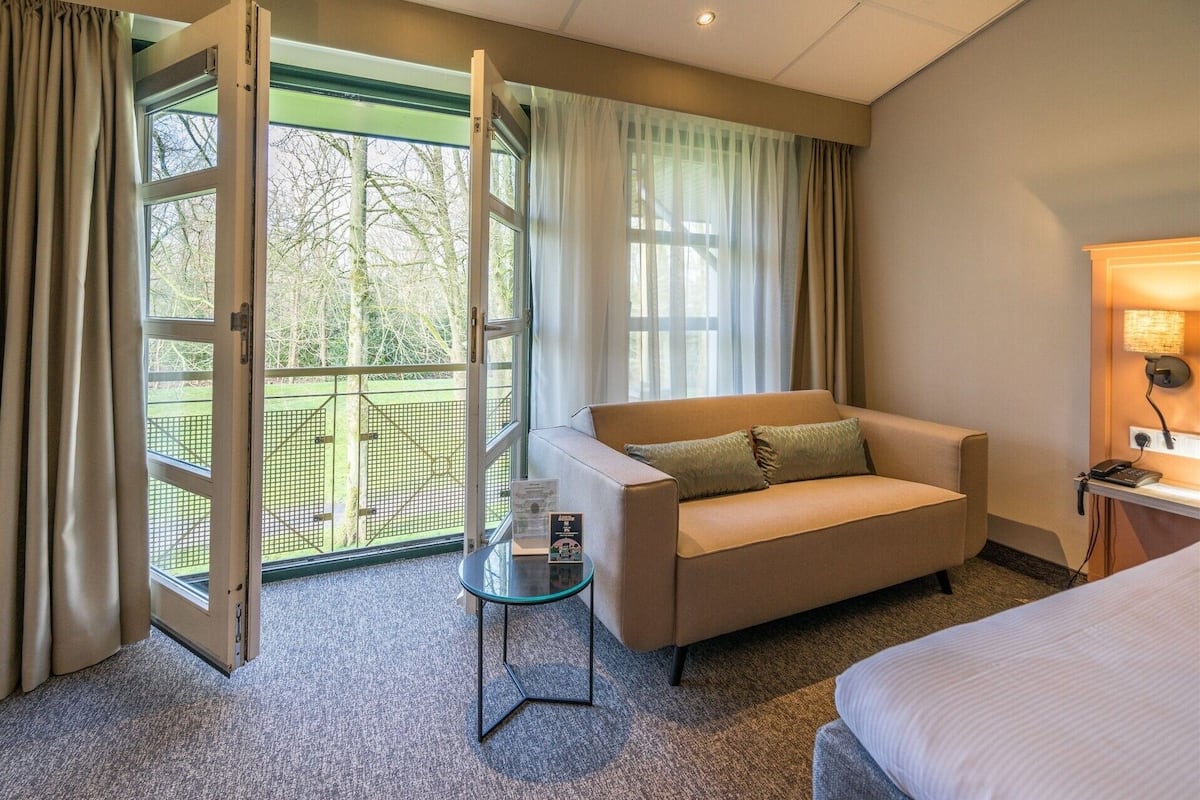 Une chambre d’hôtel avec un canapé, une petite table, un lit, un bureau avec une lampe et une vue sur un espace extérieur verdoyant, visible à travers une porte vitrée.