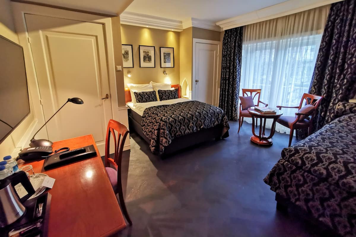Une chambre d’hôtel avec un lit, un bureau, une chaise, une table et une fenêtre avec des rideaux.