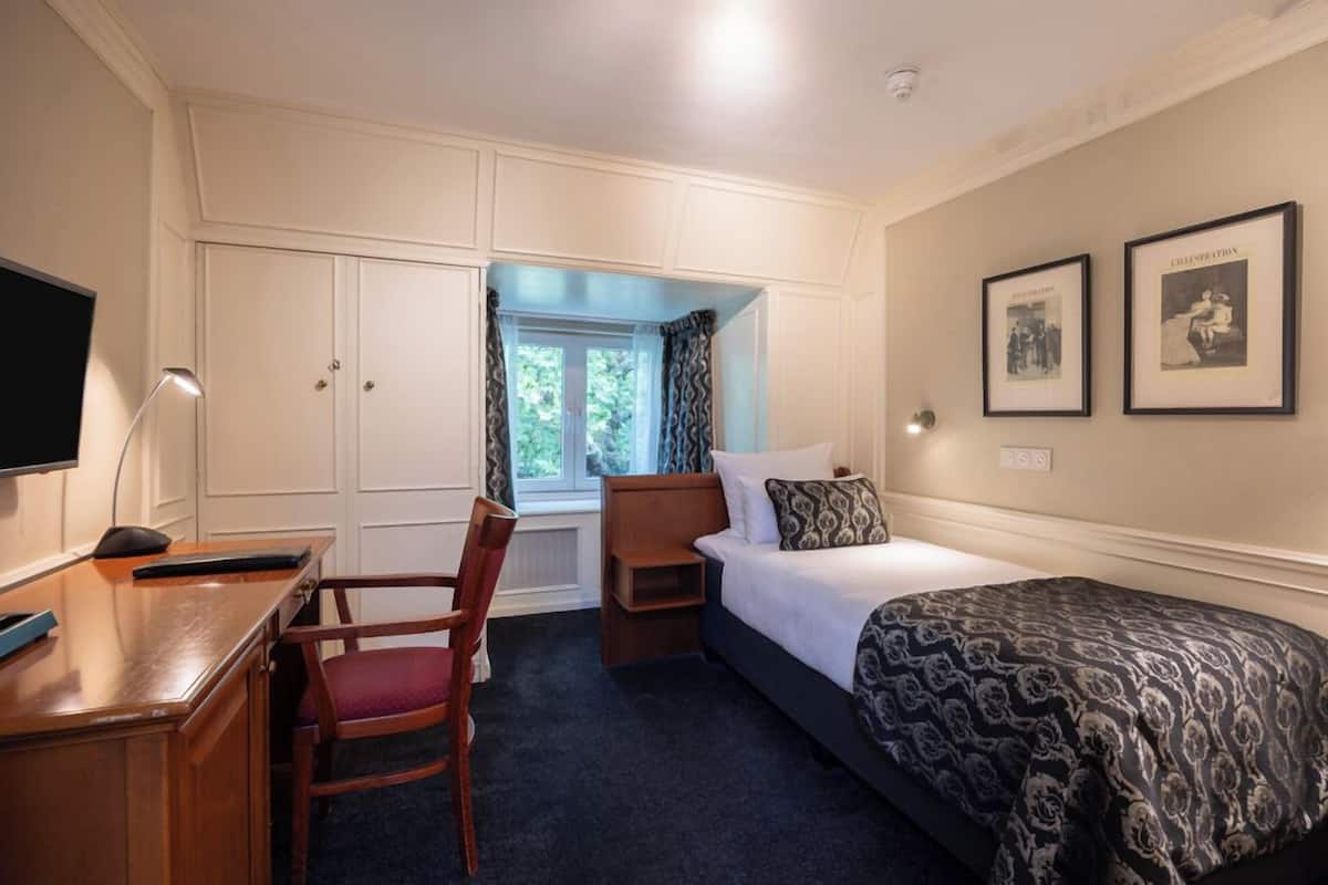 Une chambre d’hôtel avec un lit, un bureau, une chaise, une armoire et des tableaux encadrés au mur.