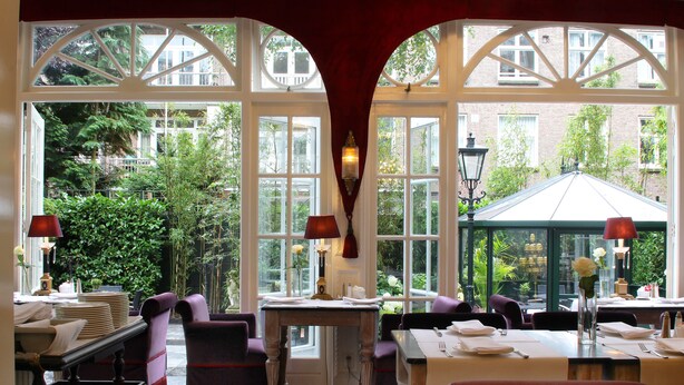 Un espace repas avec des tables dressées, de grandes fenêtres offrant une vue sur la verdure et les bâtiments, et une arche rouge encadrant les fenêtres.