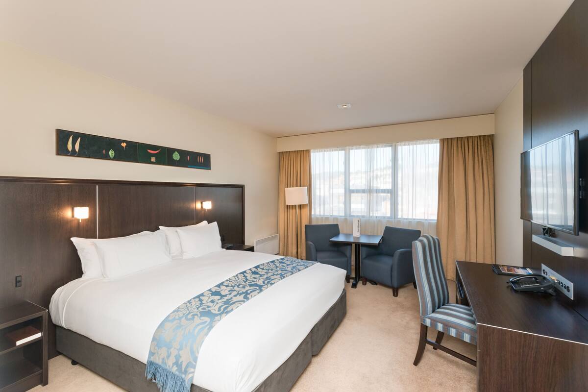 A hotel room with a large bed, a desk, a chair, a TV, and a window with curtains.