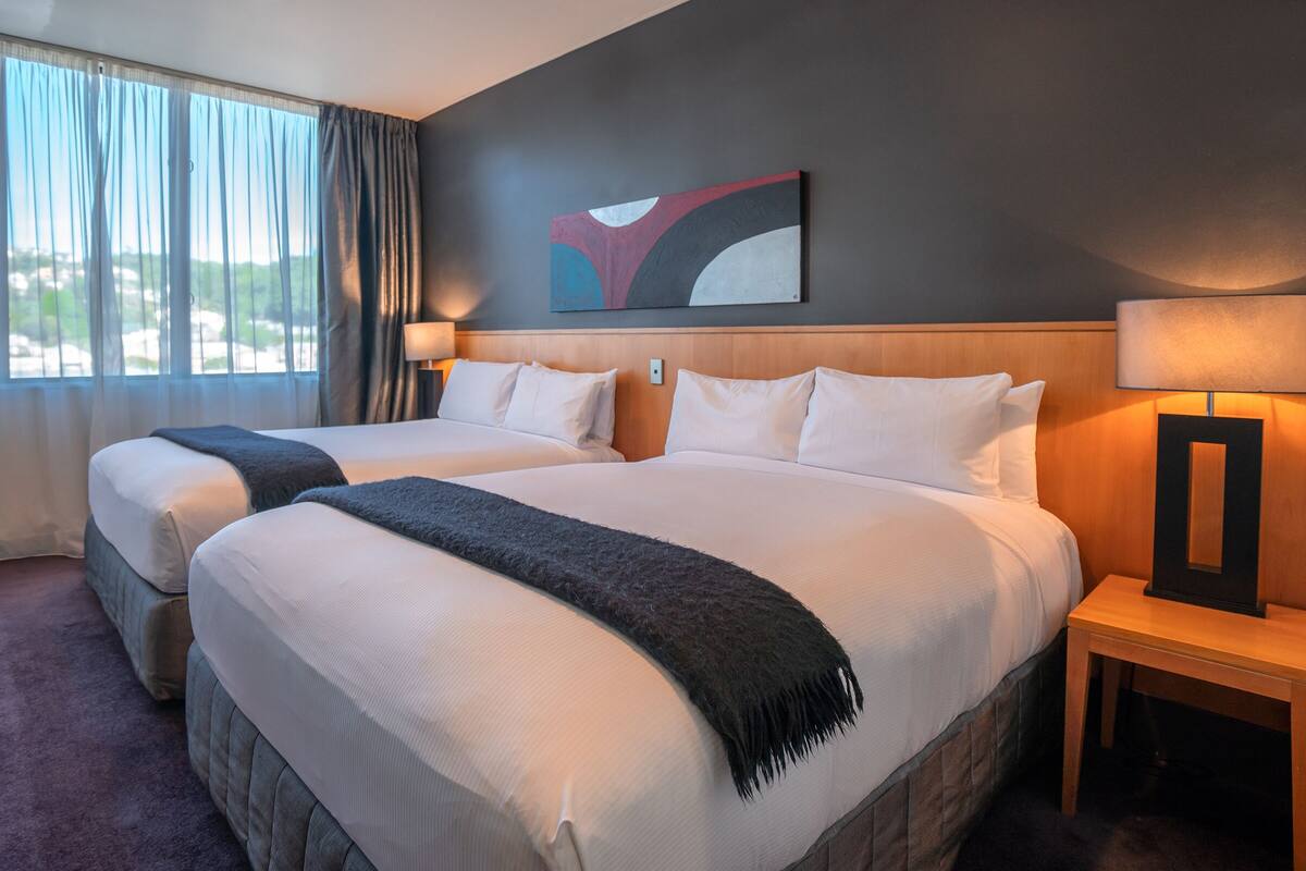 A hotel room with two beds, a large window with curtains, a painting on the wall, and a nightstand with a lamp.