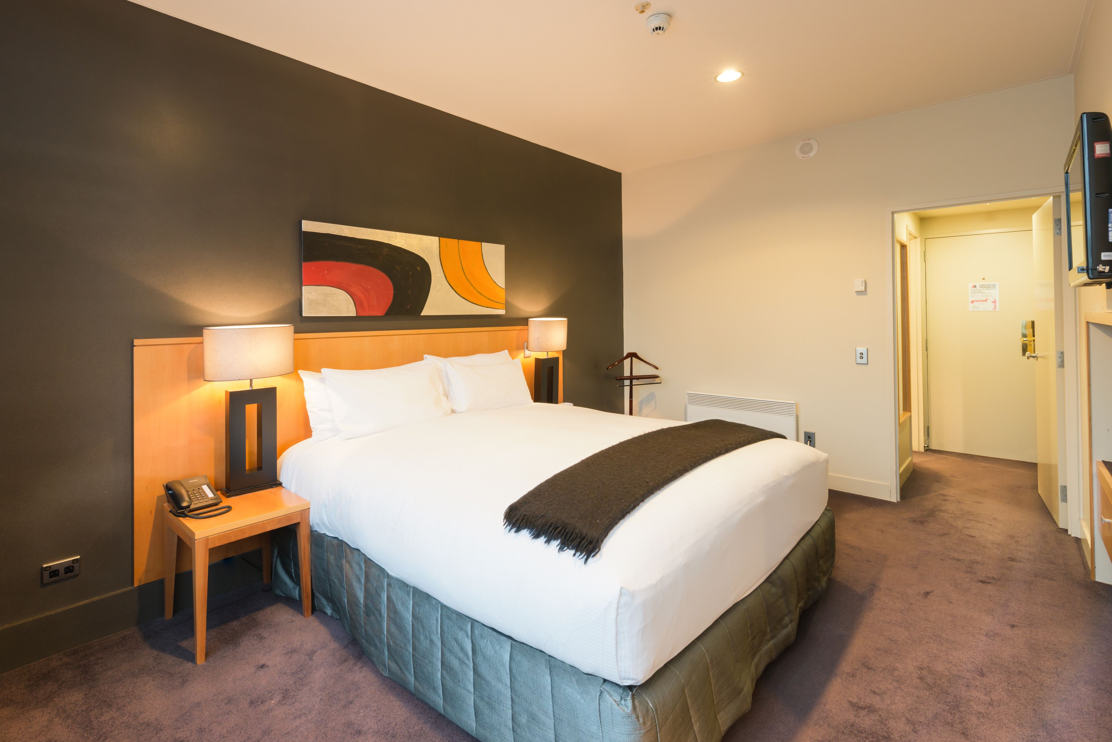A hotel room with a large bed, bedside lamps, a telephone, a painting on the wall, and a door leading to another room.