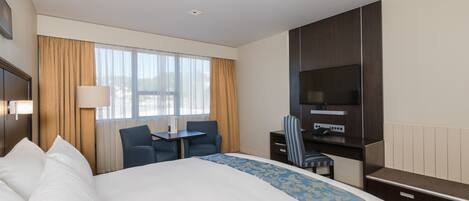 A hotel room with a large bed, a desk with a chair, a television, and a window with curtains.