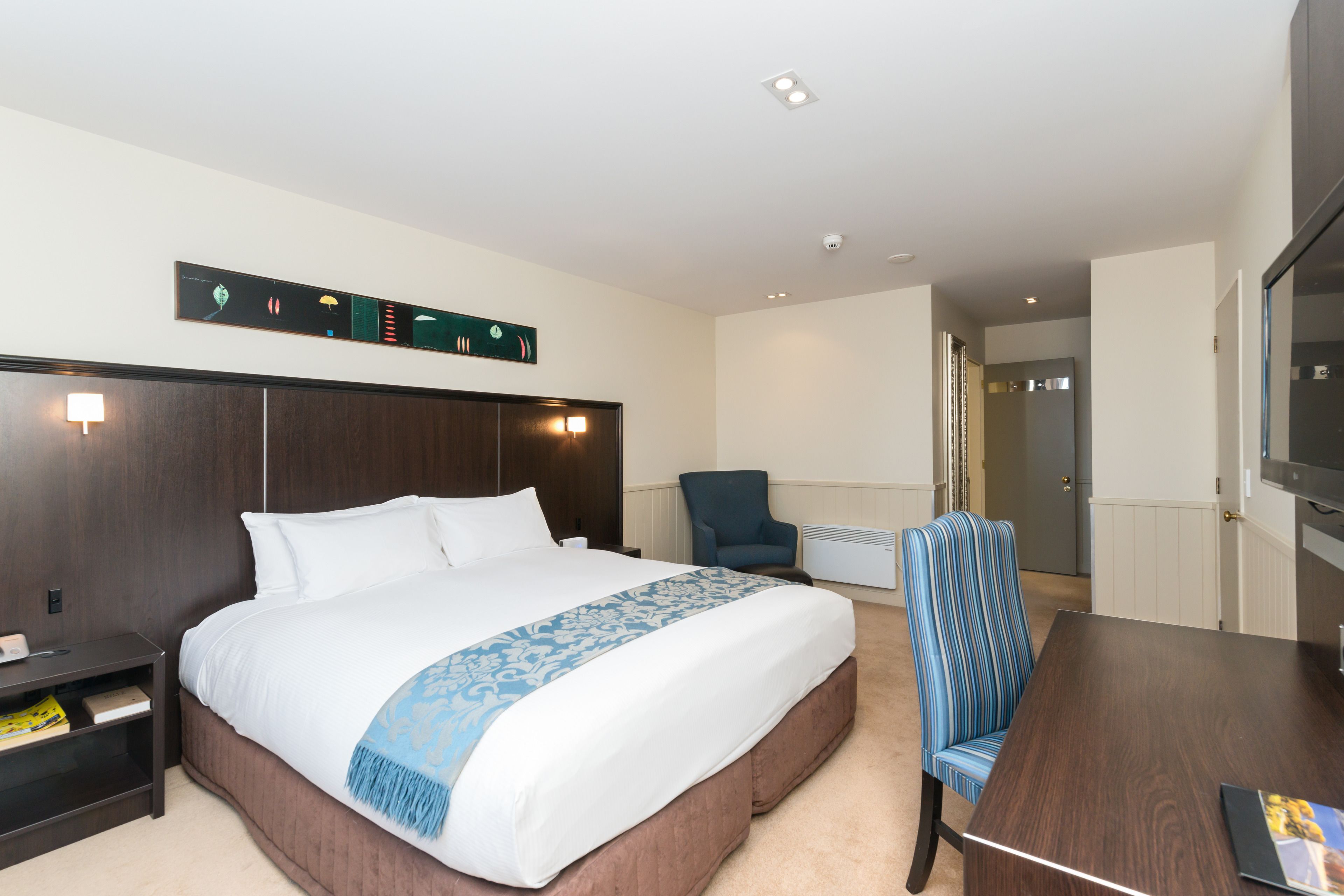 A hotel room with a large bed, a wooden headboard, a blue armchair, a wooden dining table, and a wall-mounted light fixture.