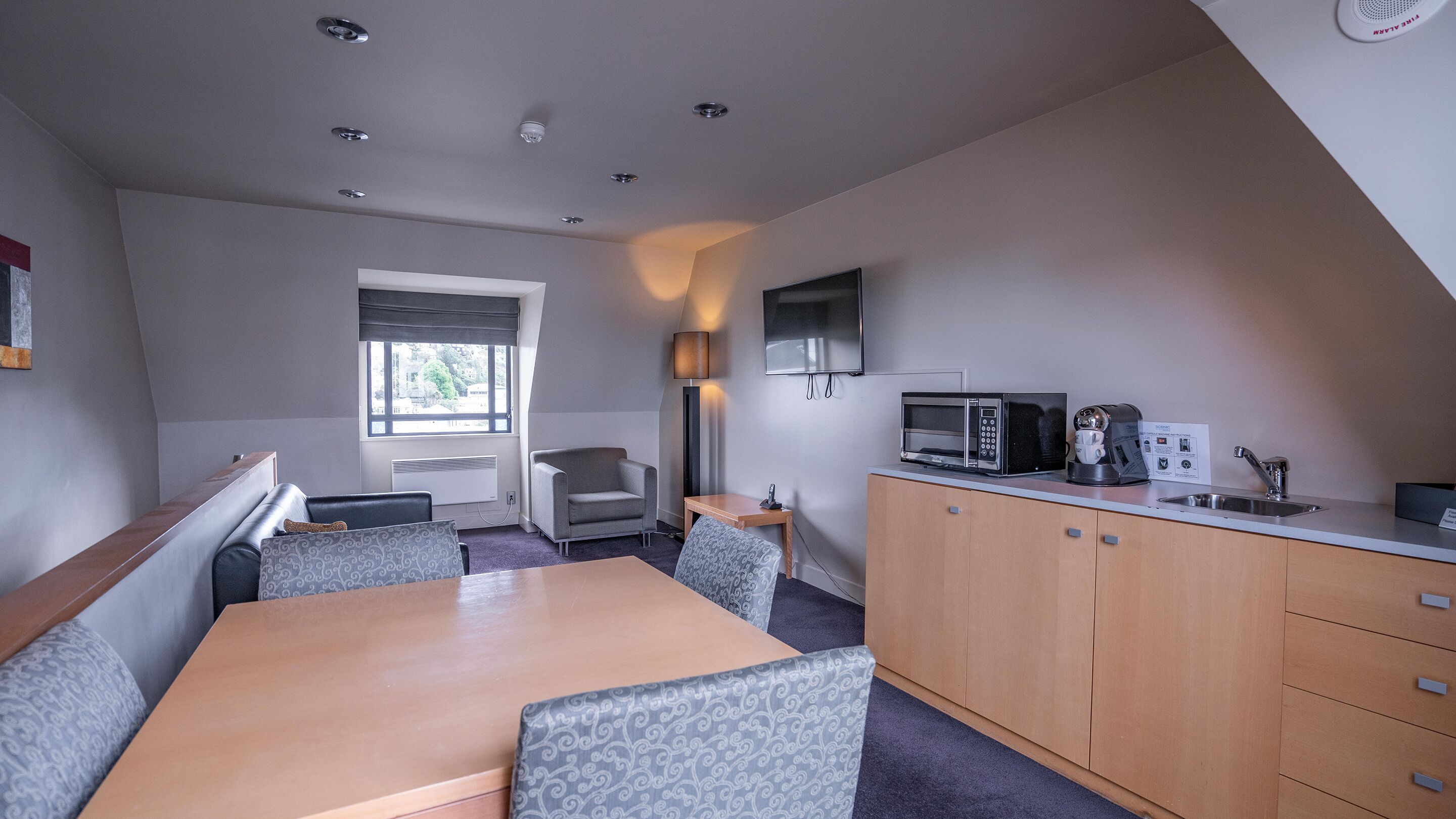 A compact hotel room with a kitchenette, a dining area, a seating area, and a window providing a view of the outdoors.