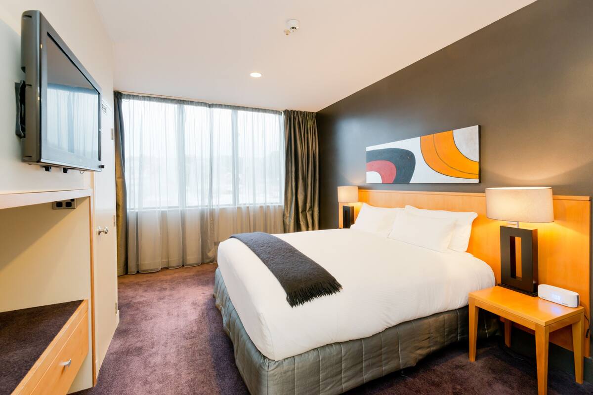 A hotel room with a bed, a TV mounted on the wall, a bedside table with a lamp, and a window with curtains.