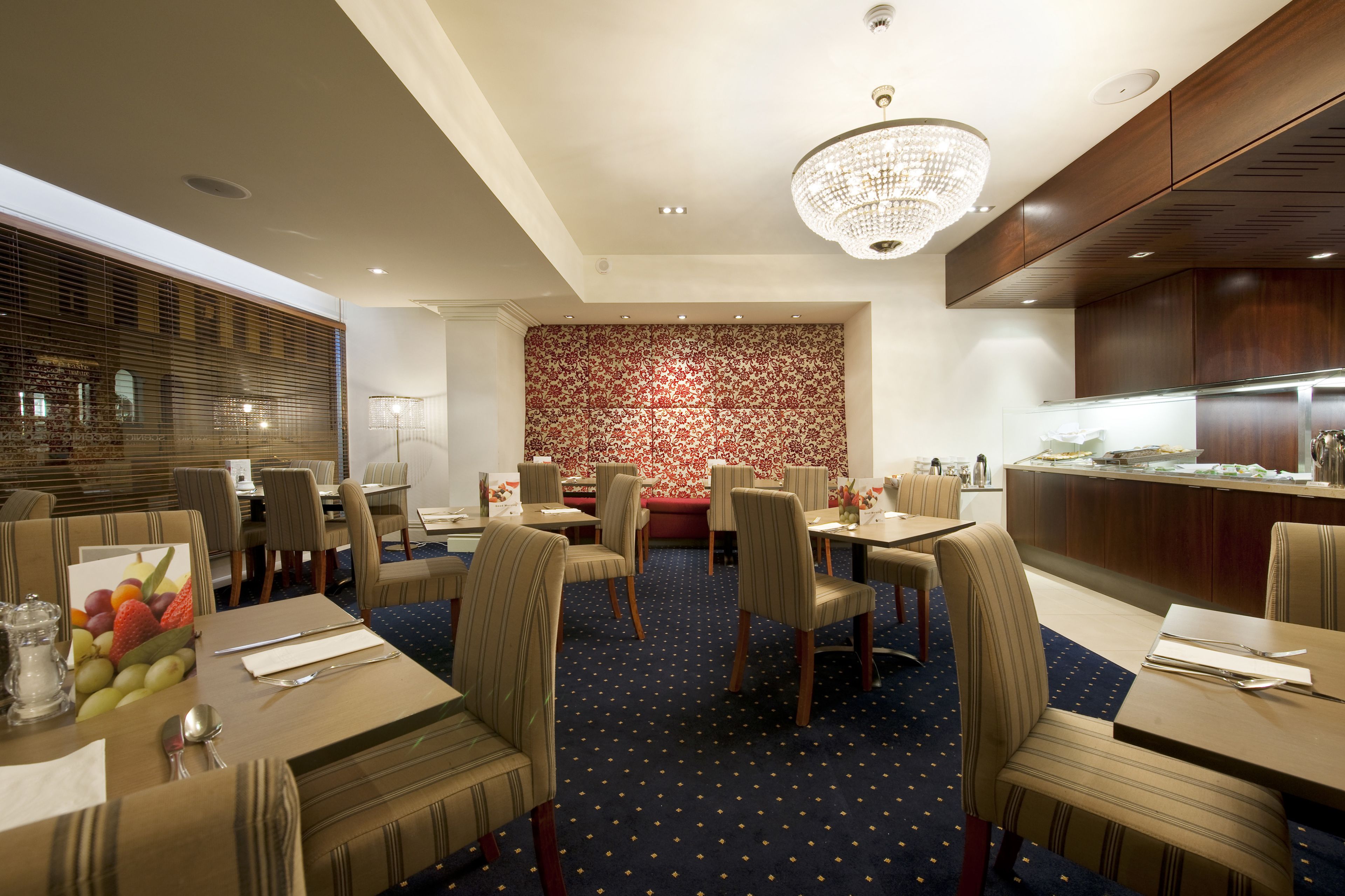 A hotel dining area with tables, chairs, a chandelier, and a patterned wall.