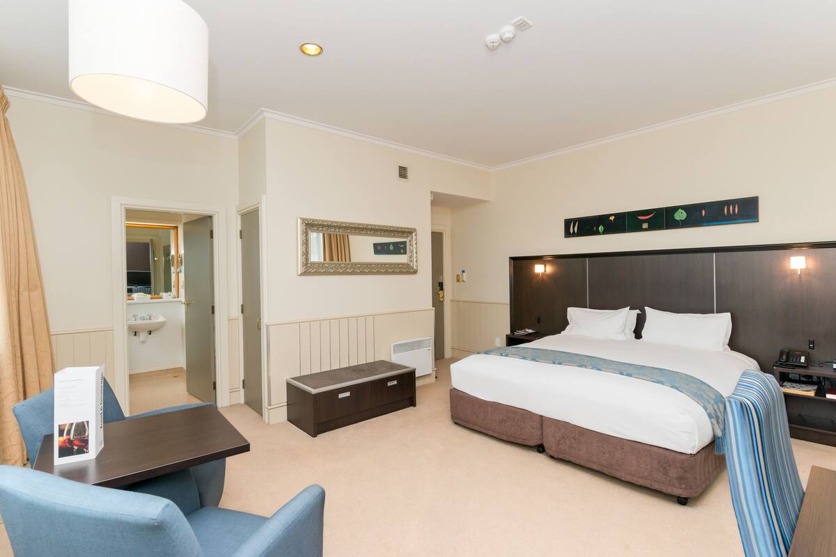 A hotel room with a large bed, a bedside table, a chair, a small desk, a mirror, a nightstand, and a wall-mounted light fixture.