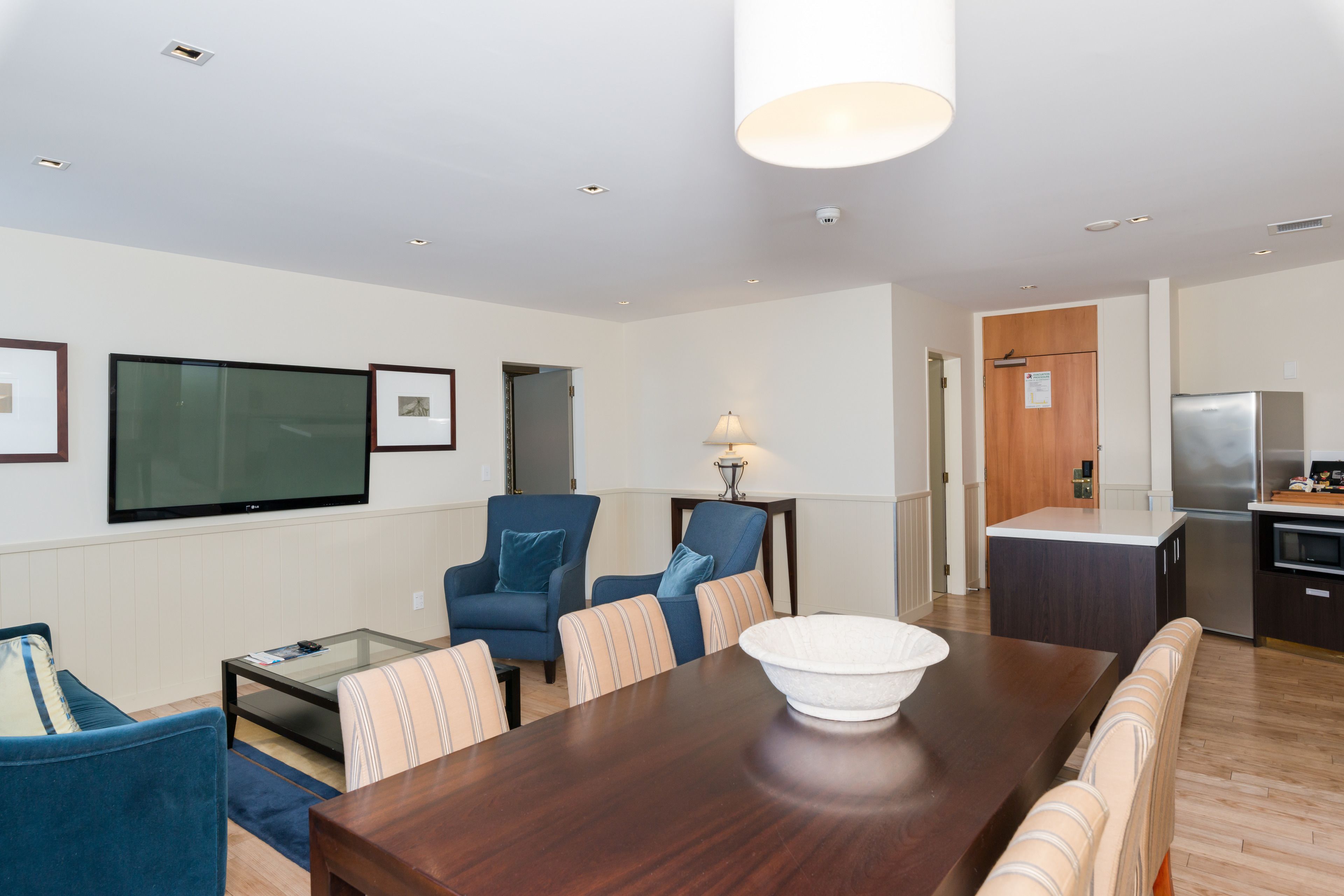 A modern living room with a dining table, blue armchairs, a glass coffee table, and a kitchen area with a refrigerator and stove.