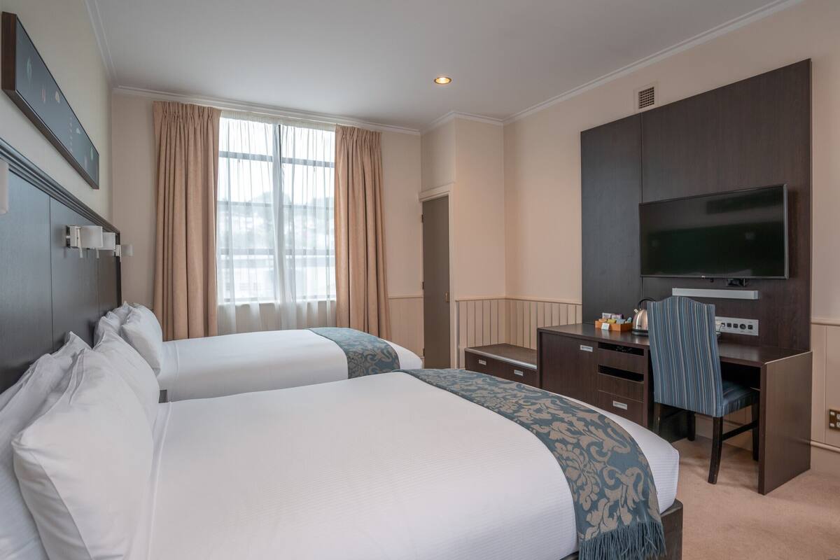 A hotel room with two beds, a desk, a television, and a window with curtains.
