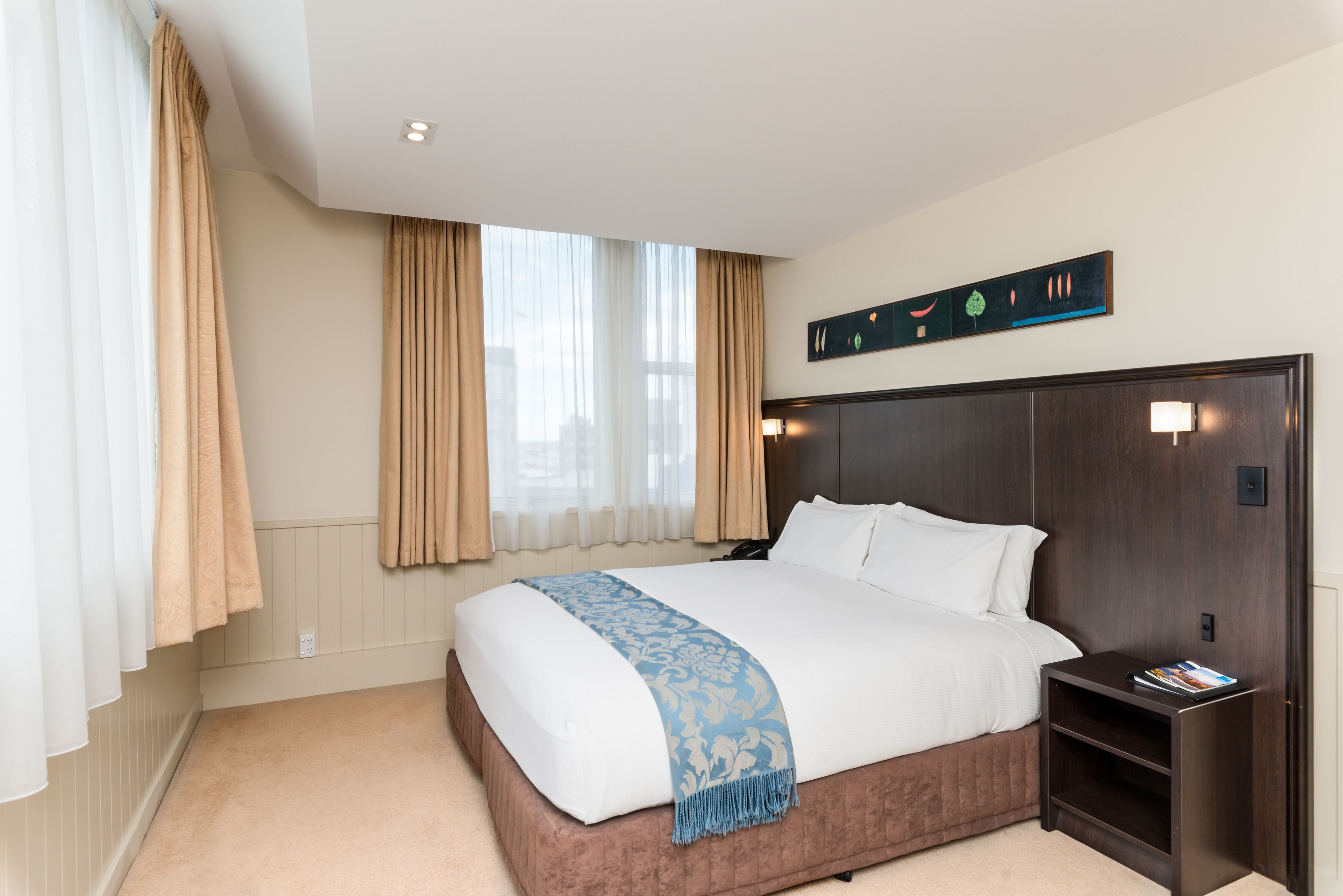 Hotelkamer met een groot bed, beige gordijnen, een houten hoofdeinde, een nachtkastje met een tijdschrift, en een kunstwerk aan de muur.