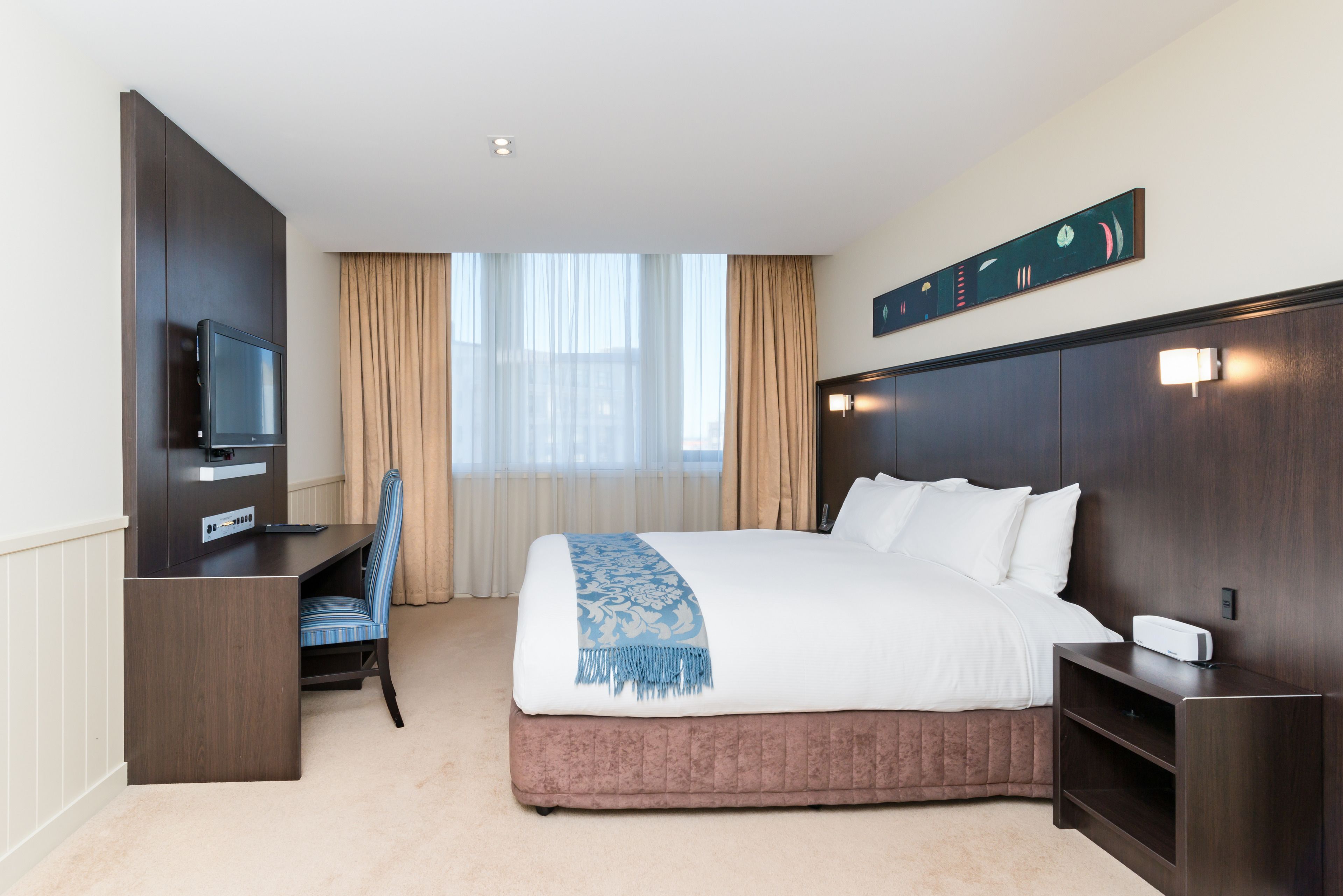 Une chambre d’hôtel avec un lit, un bureau avec une télévision, une chaise et une fenêtre avec des rideaux.