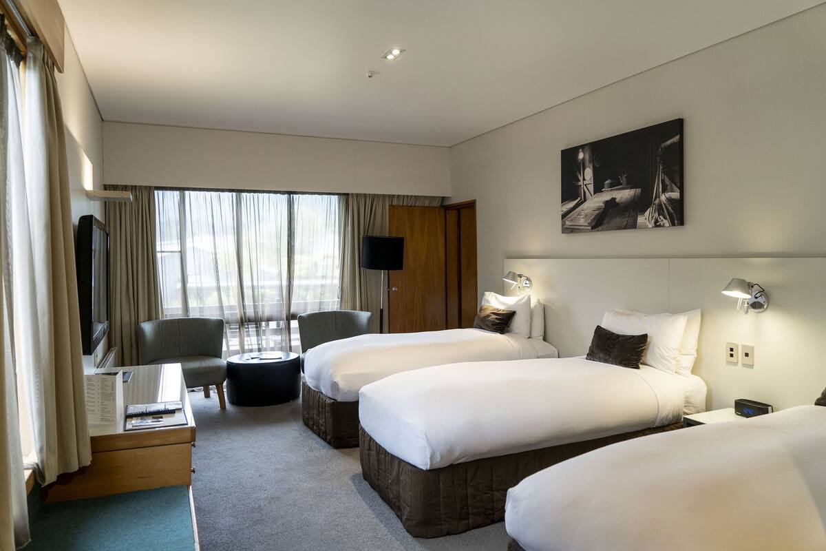 Una habitación de hotel con dos camas, un escritorio, una silla, un televisor y un amplio ventanal con cortinas.